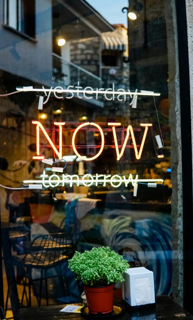 Colorful neon sign on a glass storefront with urban reflections and greenery.