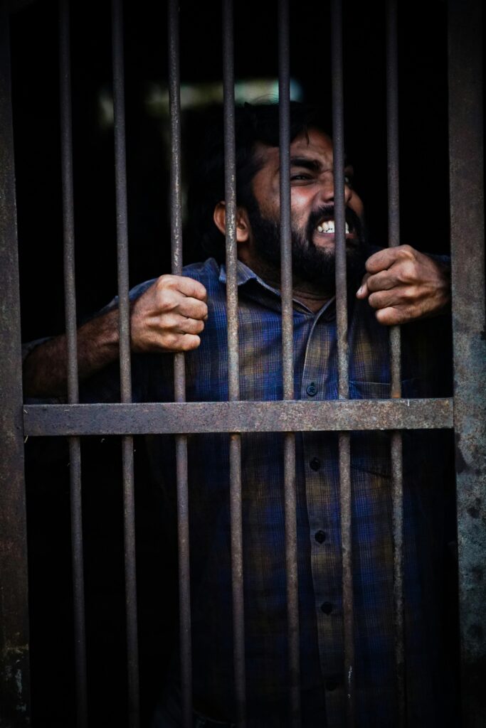 A bearded man showing intense emotion while gripping prison bars, conveying confinement and struggle.