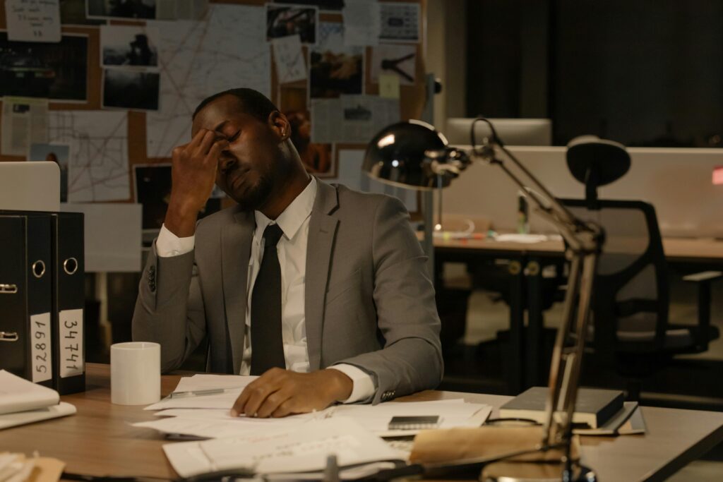 A weary man in a suit sits in an office, surrounded by paperwork, looking exhausted.