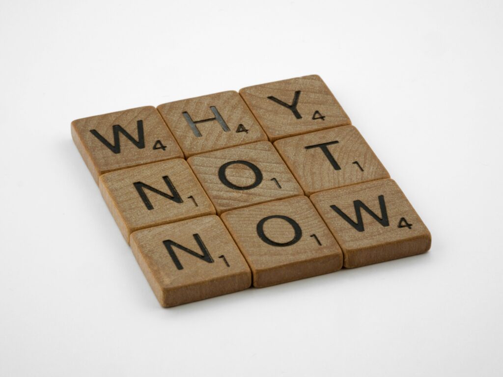 Wooden letter tiles form the motivating phrase 'Why Not Now' on a white background, encouraging action and decision-making.