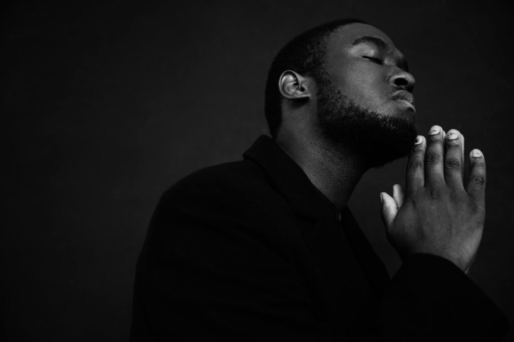 pexels-photo-7533332-7533332 Monochrome handsome unshaven African American male in black suit praying with hands clasped under chin