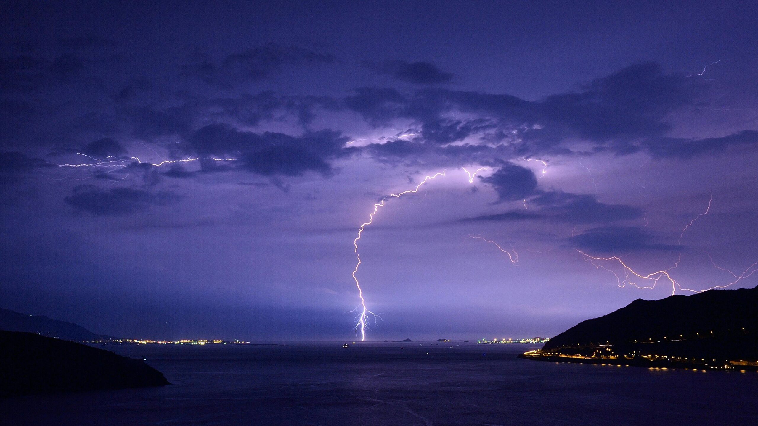 Spectacular lightning strikes illuminate a nighttime ocean view with dramatic clouds and city lights.
