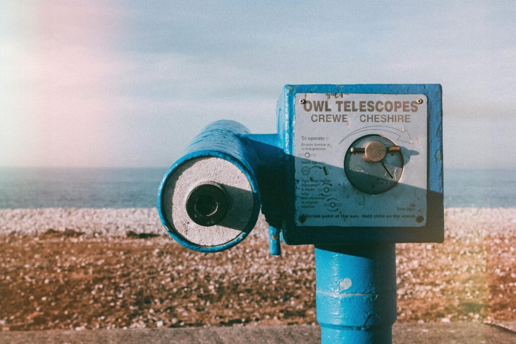 Vintage blue telescope by a pebble beach with ocean view under a clear sky.