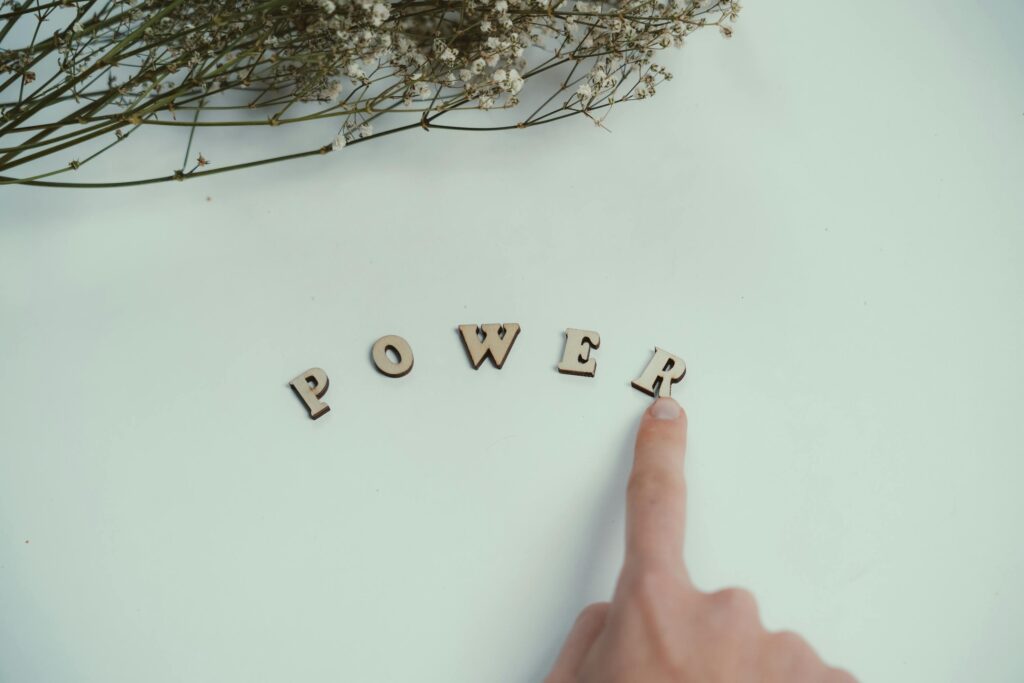 Finger points at wooden letters spelling 'Power' beside delicate flowers on a light background.