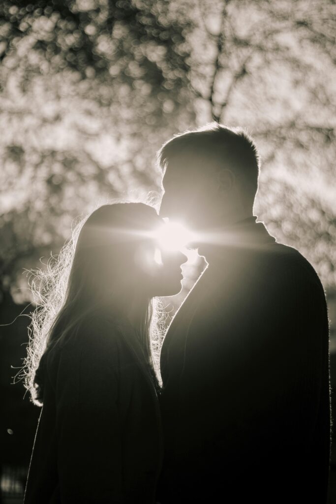 A couple shares a tender kiss in a backlit silhouette, creating a romantic sunset scene outdoors.