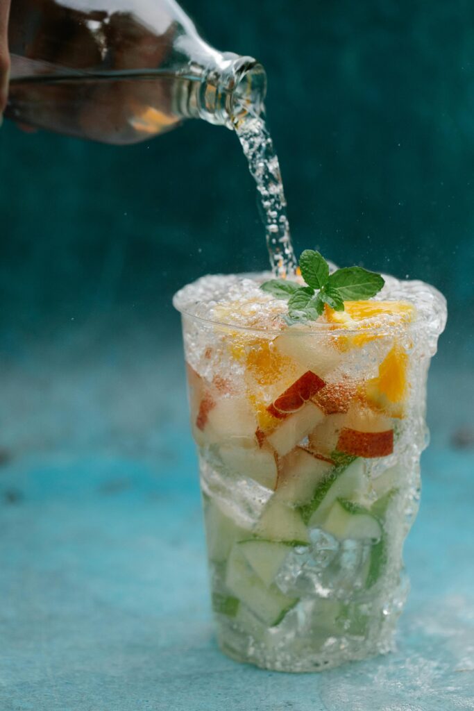 A vertical shot of sparkling water being poured over a mix of fresh fruits and mint on a blue background.